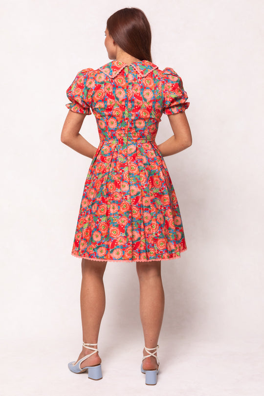 A woman faces away in the Ivy City Co Sophia Short Dress in Poppy, a short, colorful poppy print dress with puffed sleeves and a fitted waist. She has long brown hair and wears light blue heeled sandals with ankle straps.