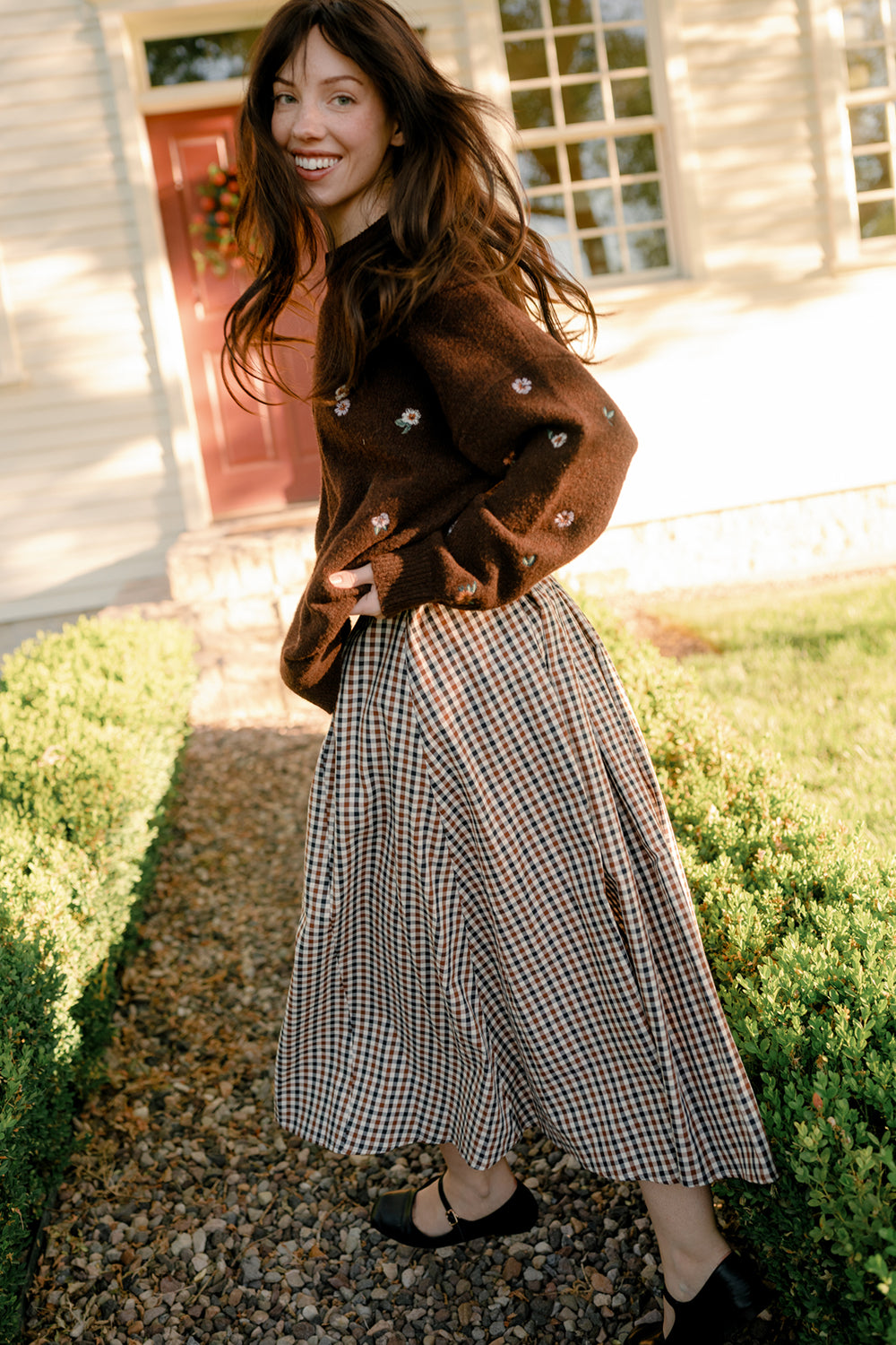 A woman with long brown hair smiles as she walks outside on a gravel path, dressed in a brown sweater adorned with small flowers and the Ivy City Co Sophia Skirt in Chestnut Gingham—a charming cotton skirt with pockets. She is near bushes and a light-colored house with large windows.