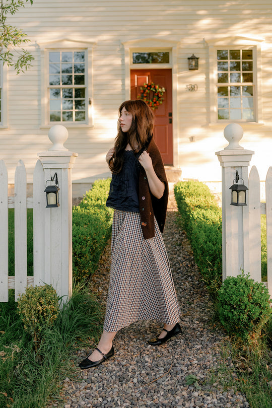 A woman wearing the Ivy City Co Sophia Skirt in Chestnut Gingham with pockets, paired with a black top and brown cardigan, stands on a stone path in front of a white house with a red door and white picket fence on a sunny day.