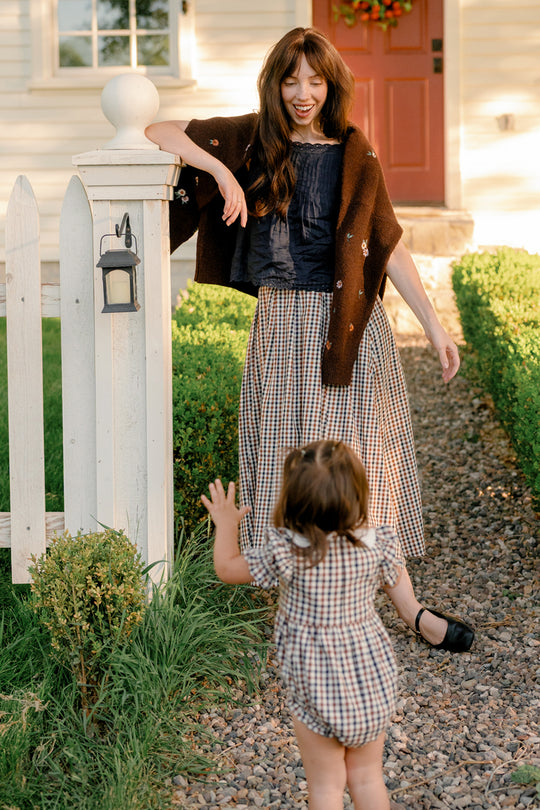 Wearing the Ivy City Co Sophia Skirt in Chestnut Gingham, a woman smiles by a white fence as a young child in plaid runs toward her along a stone path, with a house and red door visible in the background.