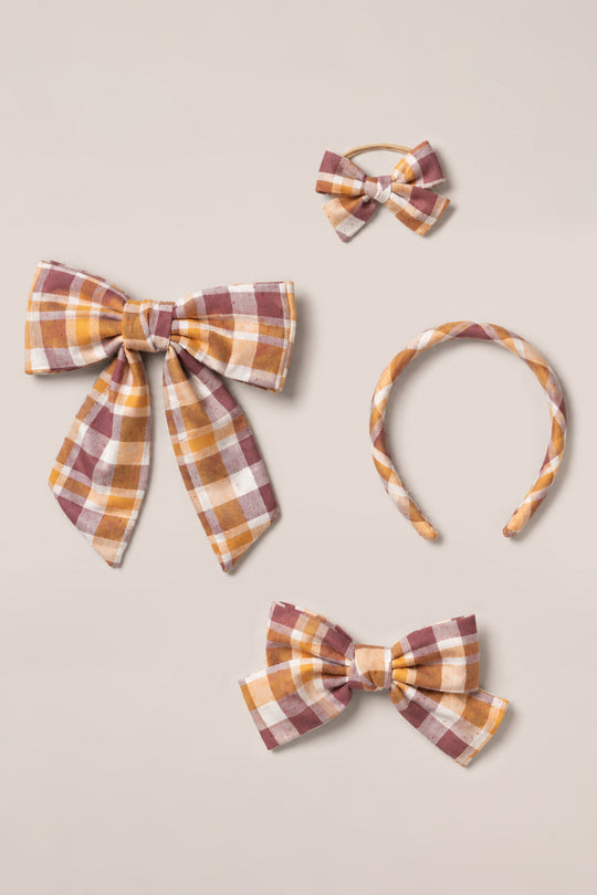 A set of four matching “Hair Bows in Spiced Plaid” by Ivy City Co—featuring a large cotton hair bow, a smaller bow on a clip, a bow on an elastic band, and a fabric-covered headband in mustard, burgundy, and cream plaid—arranged on a light beige background.