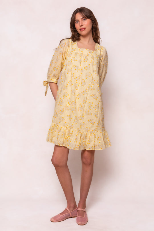 A woman stands against a light background in Ivy City Cos Sunny Dress in Yellow Floral, a loose-fitting, maternity-friendly swiss dot cotton dress with elbow-length sleeves and ruffled hem. She pairs it with pink Mary Jane shoes, looking forward with one hand in her pocket.