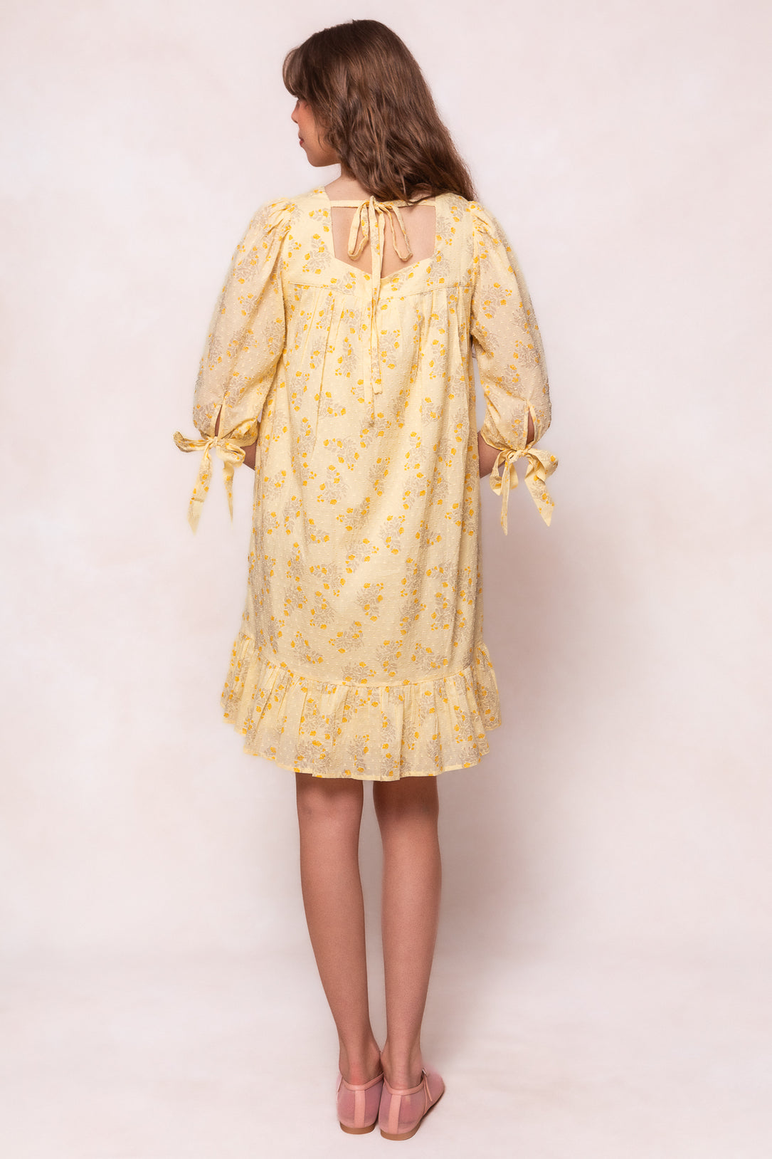A woman with brown hair faces away, wearing the Ivy City Co Sunny Dress in Yellow Floral—a maternity-friendly style with puff sleeves tied at the elbows and a ruffled hem—paired with pink shoes against a soft, neutral background.