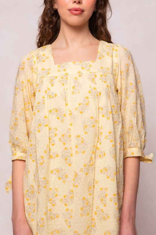 A woman wears the Sunny Dress in Yellow Floral by Ivy City Co, a maternity-friendly swiss dot cotton dress with a square neckline, elbow-length sleeves, decorative ties, loose fit, and small yellow floral print.