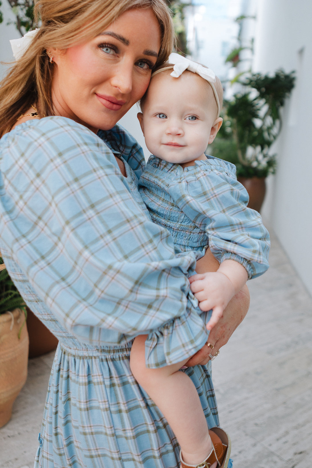 A woman holds a smiling baby wearing the Ivy City Co Baby Madeline Romper in Light Blue Plaid with a white bow headband and smocked bodice; both are dressed in matching outfits as they stand outdoors near green plants.
