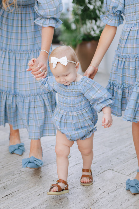 A baby in the Ivy City Co Baby Madeline Romper in Light Blue Plaid, with a smocked bodice and white bow, walks hand-in-hand with two adults in matching blue plaid dresses. The bright scene features a blurred plant background.