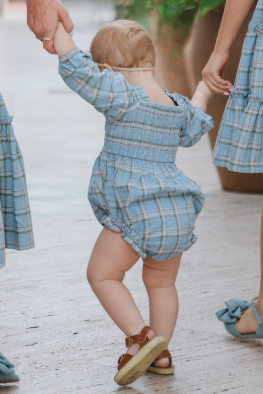 A toddler wears the Ivy City Co Baby Madeline Romper in Light Blue Plaid, holding hands with two adults in matching blue plaid outfits. The toddler steps forward in brown sandals, framed by the adults on each side.