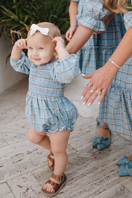 A smiling baby in Ivy City Co’s Baby Madeline Romper in Light Blue Plaid, featuring a smocked bodice and white bow, stands holding an adult’s hands. The adult wears a matching blue plaid dress and bow sandals by greenery on stone flooring.