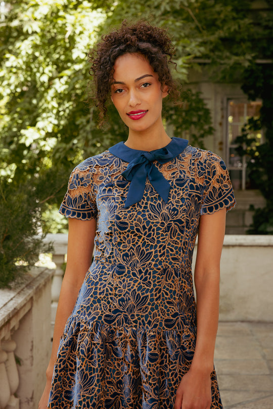 Woman wearing a patterned dress with a blue bow standing outdoors.