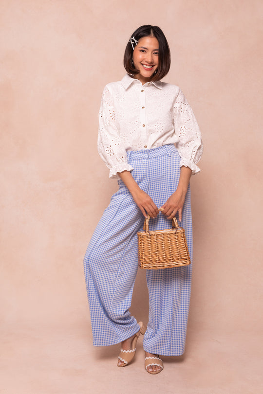 A smiling woman stands against a beige background, wearing the Ivy City Co Lane Blouse in White Eyelet, blue and white checkered wide-leg pants, white sandals, and holding a wicker handbag. Her bob is styled with a hair clip.