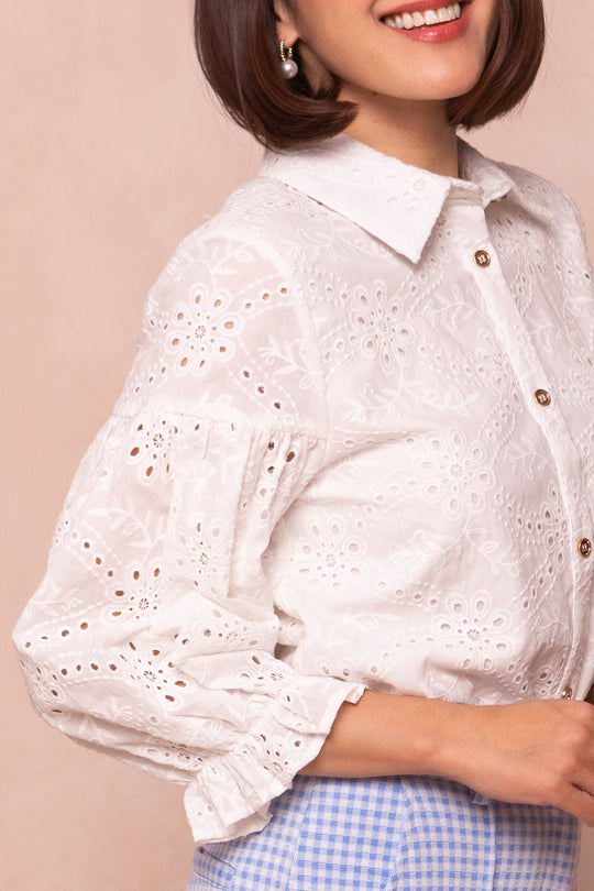 A woman wears the Lane Blouse in White Eyelet by Ivy City Co, a nursing-friendly cotton blouse with puffed sleeves, styled with blue gingham pants and pearl earrings. Her chin-length hair and smile complete the charming look.