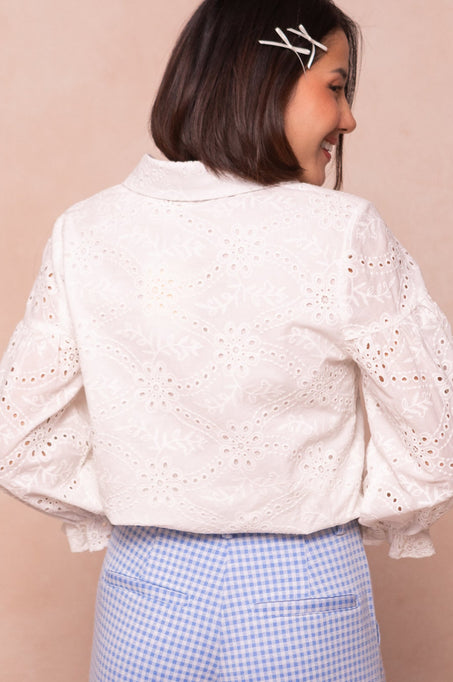 A woman with shoulder-length brown hair, wearing the Ivy City Co Lane Blouse in White Eyelet and blue gingham pants, stands smiling with her back turned. She has two white hair clips in her hair.