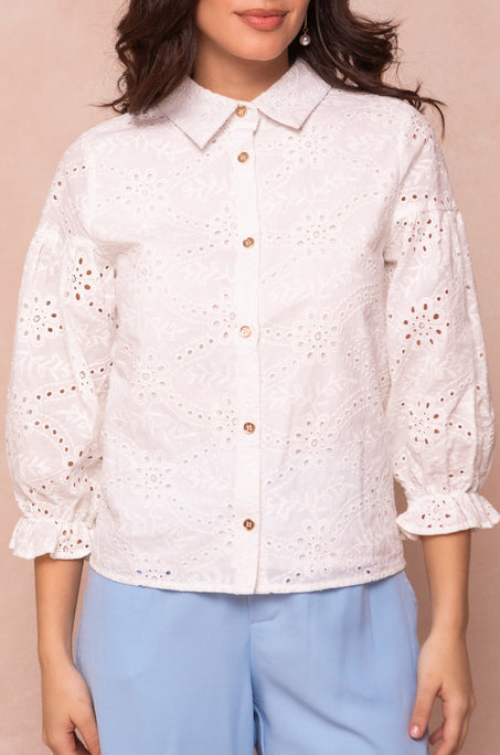 A woman wears the Lane Blouse in White Eyelet by Ivy City Co, featuring a collared, button-up front, puffed sleeves, and ruffled cuffs. She pairs it with light blue pants; her face is partially out of frame.