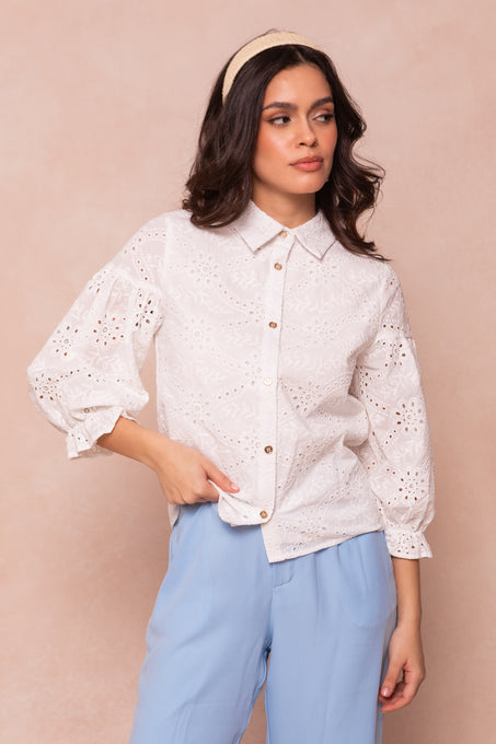 A woman with wavy dark hair, a beige headband, and light blue pants wears the Lane Blouse in White Eyelet by Ivy City Co, featuring balloon sleeves, while standing against a soft beige background.