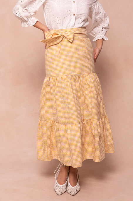 A person wears the Ivy City Co Jodi Skirt in Yellow Gingham with a white eyelet blouse and white pointed-toe shoes, standing against a beige background.