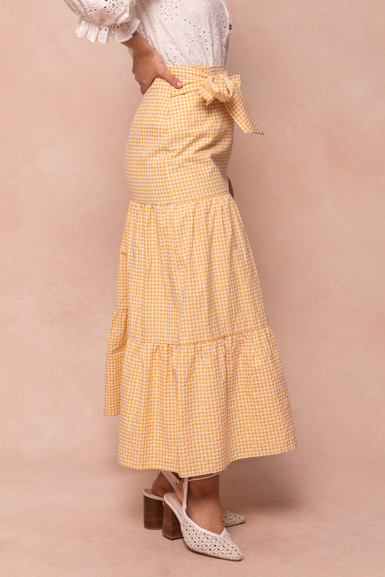 A woman wears the Ivy City Co Jodi Skirt in Yellow Gingham with a white eyelet blouse and white heeled sandals, standing against a neutral background.
