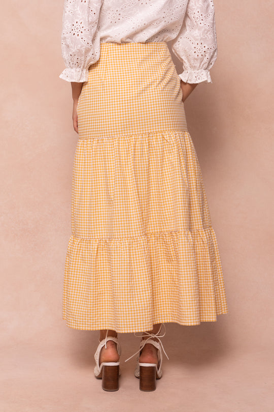 A woman stands with her back to the camera, wearing a white eyelet blouse and the Jodi Skirt in Yellow Gingham by Ivy City Co, styled with heeled sandals.