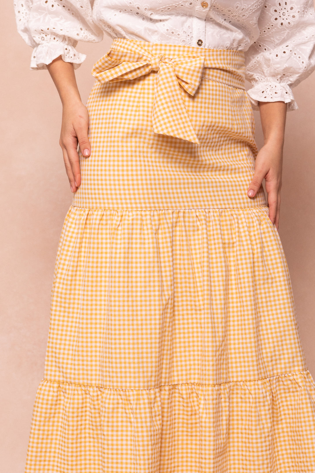 A model wears the Ivy City Co Jodi Skirt in Yellow Gingham, a cotton tiered design with a waist bow, paired with a white eyelet blouse, shown from shoulders to ankles against a light peach backdrop.