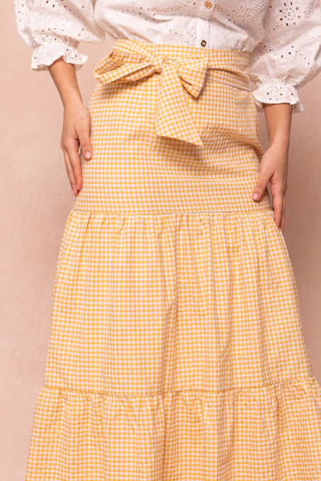 A model wears the Ivy City Co Jodi Skirt in Yellow Gingham, a cotton tiered design with a waist bow, paired with a white eyelet blouse, shown from shoulders to ankles against a light peach backdrop.