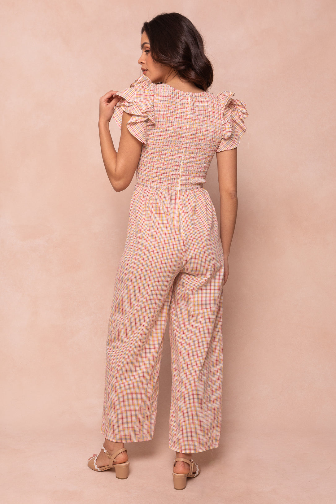 A woman with long brown hair wears the Ivy City Co Hattie Jumpsuit in Multicolor Gingham—an ankle-length style with short ruffled sleeves and wide legs. She faces away in heeled sandals against a beige background.