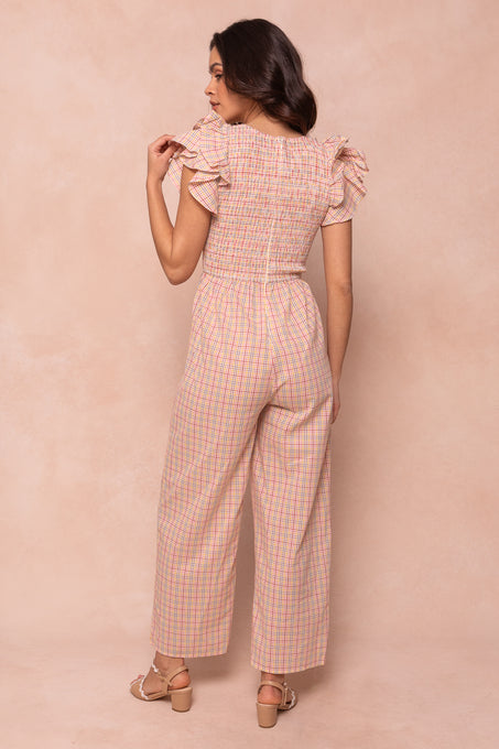 A woman with long brown hair wears the Ivy City Co Hattie Jumpsuit in Multicolor Gingham—an ankle-length style with short ruffled sleeves and wide legs. She faces away in heeled sandals against a beige background.