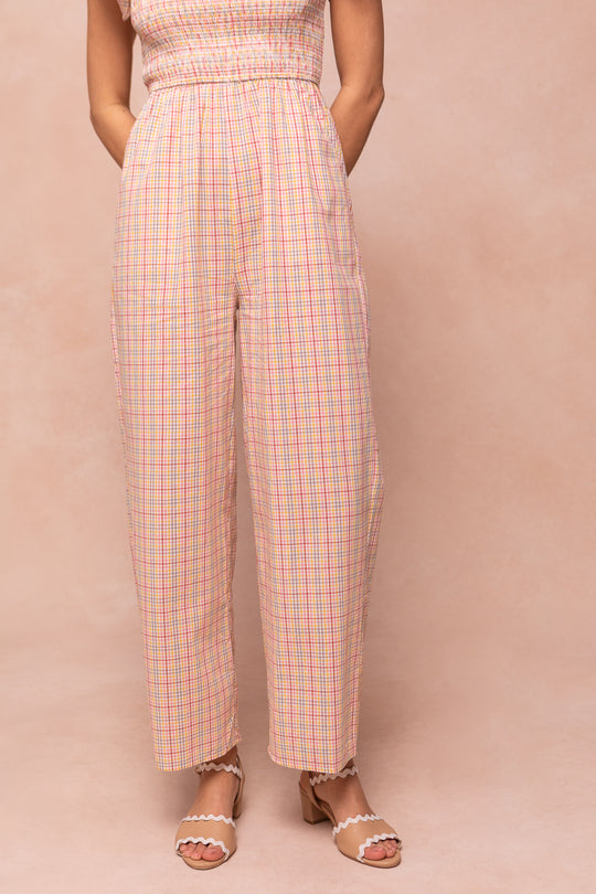 A woman models the Ivy City Co Hattie Jumpsuit in Multicolor Gingham, featuring a playful, high-waisted silhouette with pockets, paired with tan scalloped-heel sandals, set against a soft beige backdrop.