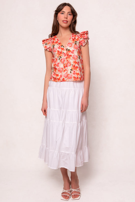 A woman with long brown hair stands against a light background, wearing the Ivy City Co Lana Blouse in Pink Floral, a tiered white midi skirt, and white strappy sandals. She has a neutral expression.