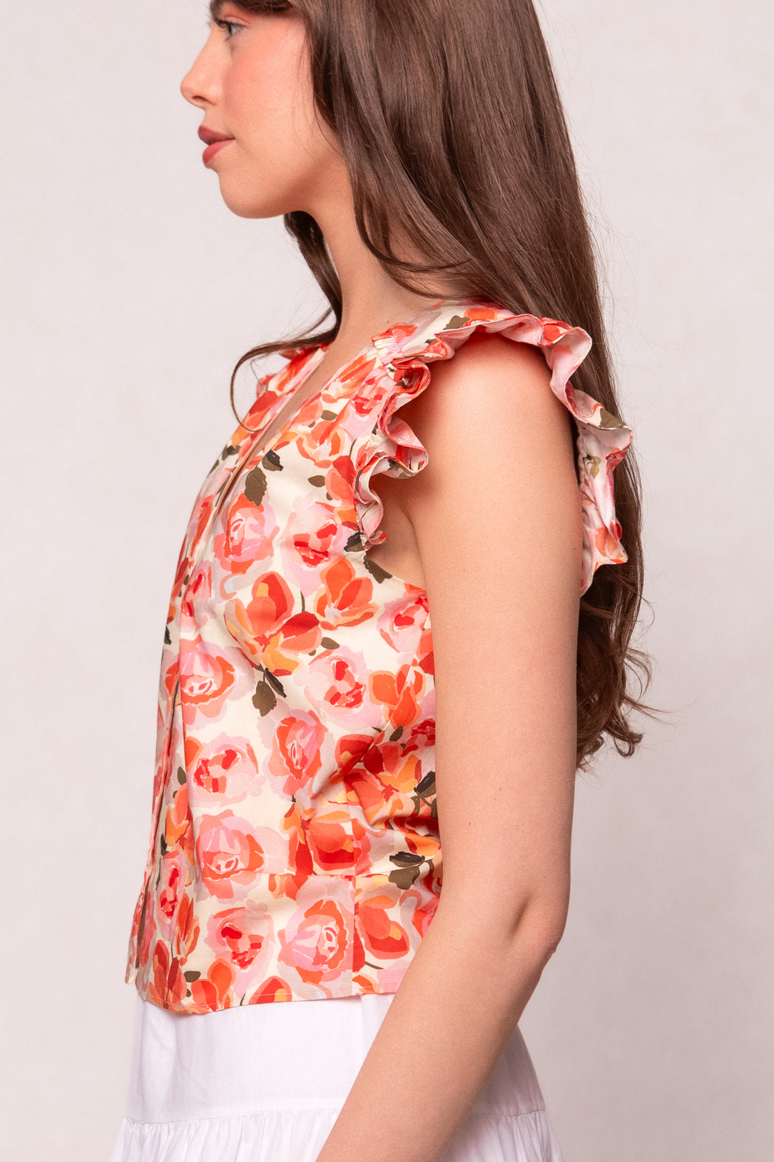 A young woman with long brown hair stands in profile against a plain background, wearing the Ivy City Co Lana Blouse in Pink Floral with ruffled shoulders and a white skirt.