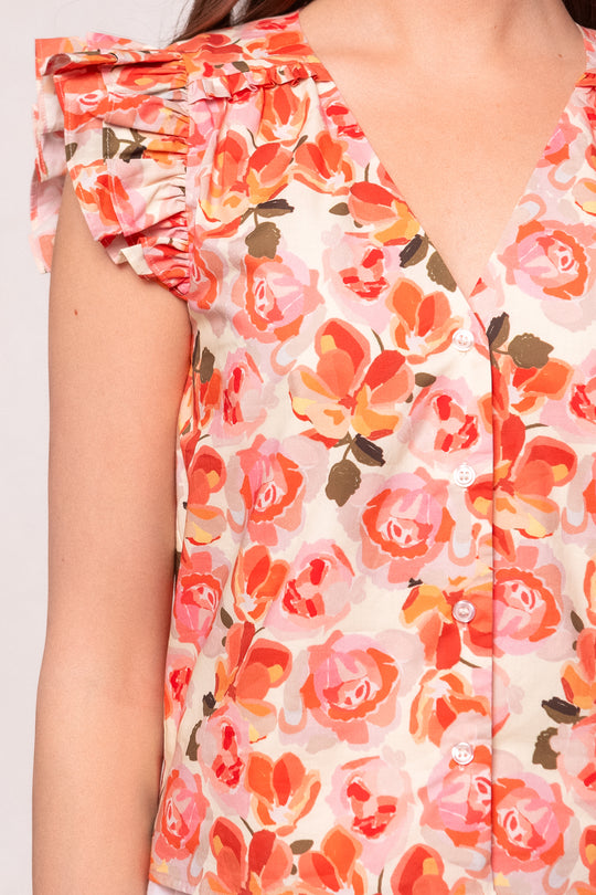 Close-up of a woman wearing the Ivy City Co Lana Blouse in Pink Floral—a cotton button-up with white buttons, ruffled shoulders, and a vivid pink, orange, and red floral pattern. Only her upper body is shown.