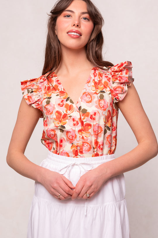 A woman wearing the Ivy City Co Lana Blouse in Pink Floral, featuring ruffled shoulders, pairs it with a white skirt and smiles against a neutral background. This cotton button-up blouse combines style and comfort.