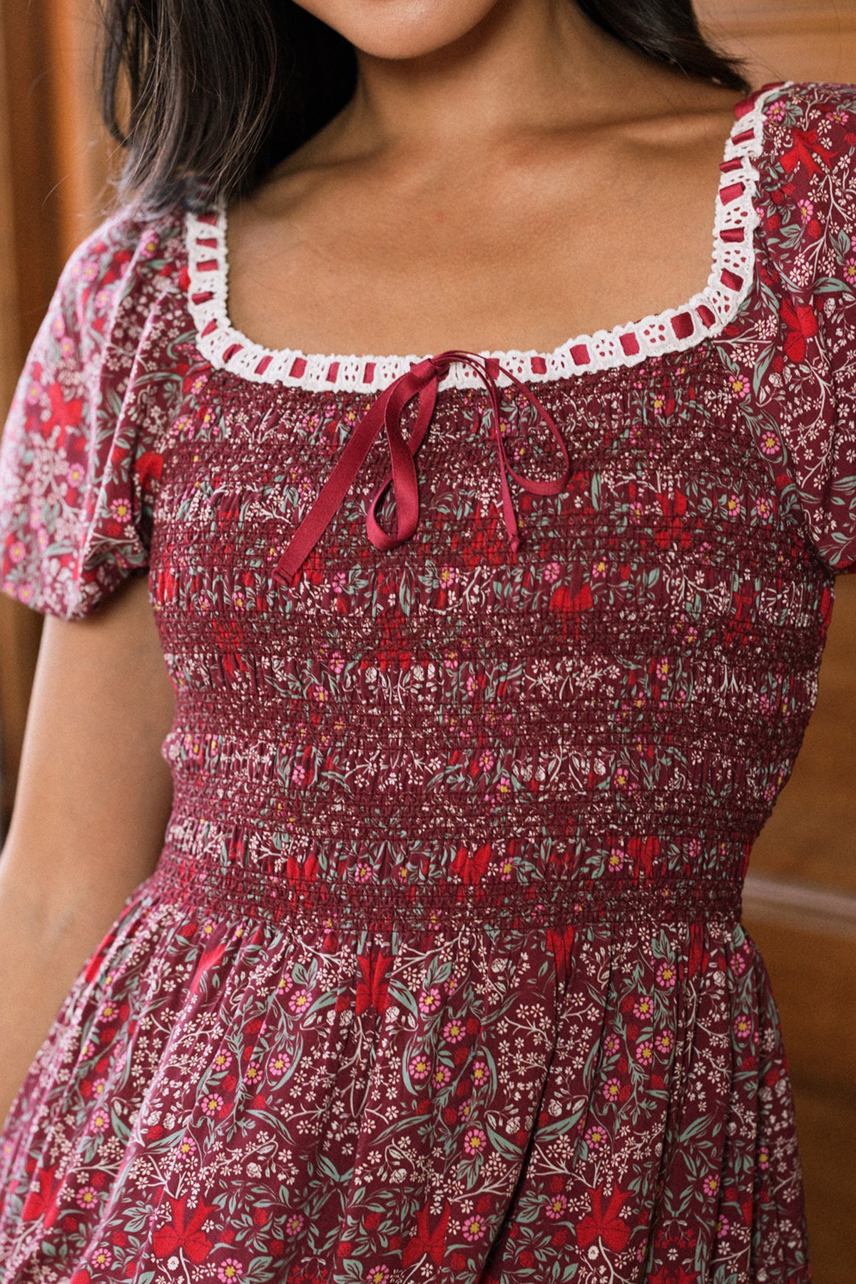 A woman wearing the Ivy City Co Sunday Best Dress in Garnet Ribboned Garden, which features a smocked bodice with puffed sleeves, a square neckline accented with white lace trim, and a small red bow at the center. The photo is cropped below her nose.