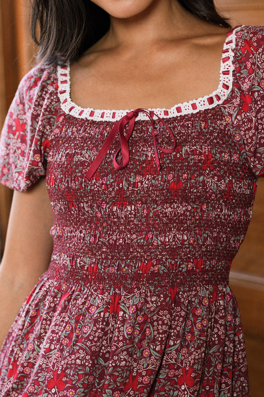 A woman wearing the Ivy City Co Sunday Best Dress in Garnet Ribboned Garden, which features a smocked bodice with puffed sleeves, a square neckline accented with white lace trim, and a small red bow at the center. The photo is cropped below her nose.