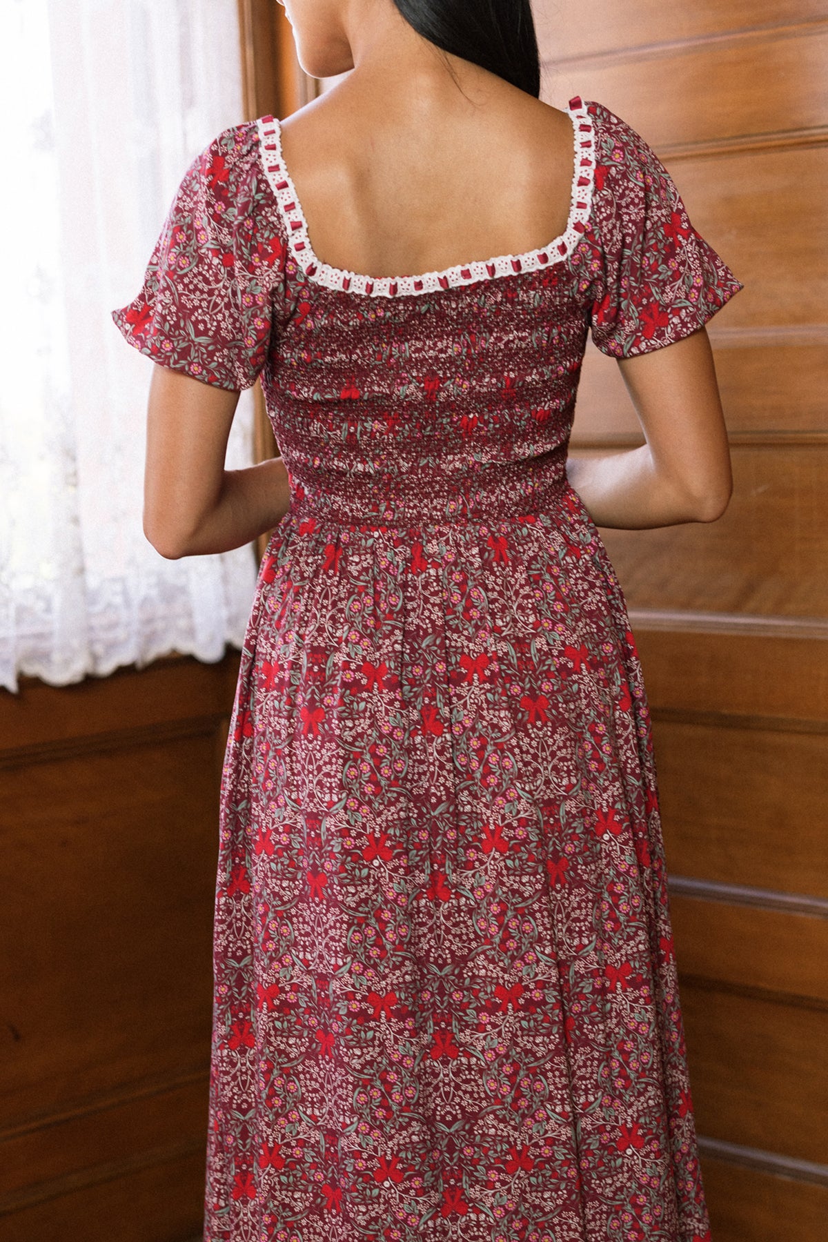 A woman stands indoors facing away, wearing the Ivy City Co Sunday Best Dress in Garnet Ribboned Garden. This floral dress features short sleeves, a square neckline adorned with white lace trim, and a charming smocked bodice—making it the perfect holiday choice as sunlight streams through a lace curtain beside her.