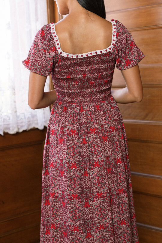 A woman stands indoors facing away, wearing the Ivy City Co Sunday Best Dress in Garnet Ribboned Garden. This floral dress features short sleeves, a square neckline adorned with white lace trim, and a charming smocked bodice—making it the perfect holiday choice as sunlight streams through a lace curtain beside her.
