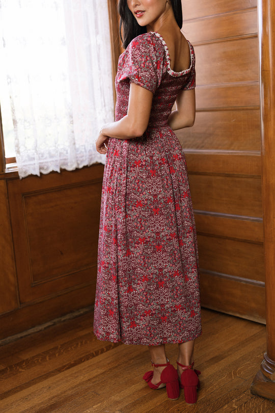 A woman in the Ivy City Co Sunday Best Dress in Garnet Ribboned Garden, featuring a smocked bodice and floral pattern, pairs her look with red heels as she poses on a wooden floor in a room with wood paneling and a window dressed with lace curtains, her head turned over her shoulder.