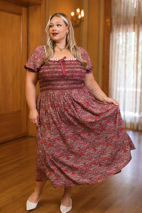 A woman with long, light hair stands indoors on a wooden floor, holding out one side of her Sunday Best Dress in Garnet Ribboned Garden by Ivy City Co. The dress features a smocked bodice and short sleeves adorned with a delicate floral pattern. She completes the look with white shoes, while elegant lighting and large windows in the background create a perfect setting for this chic holiday ensemble.