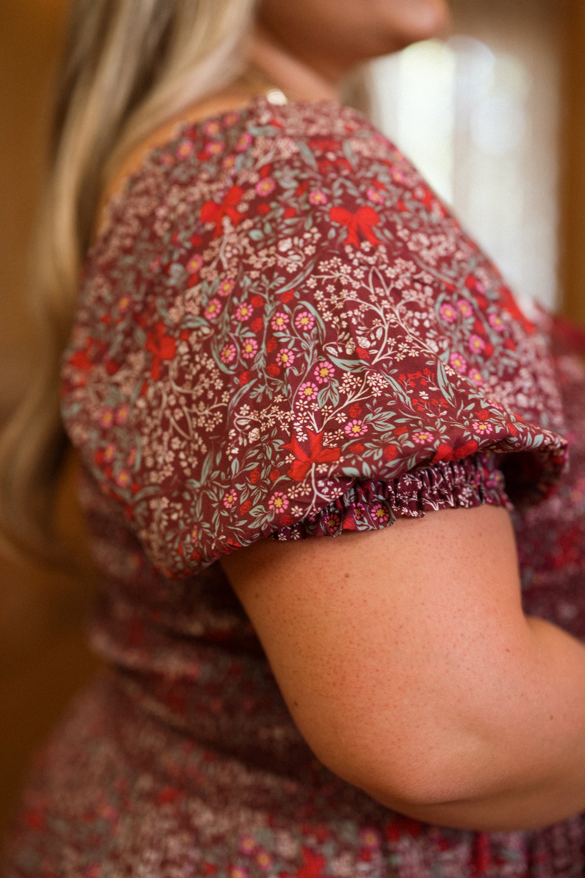 A close-up of a person wearing the Ivy City Co Sunday Best Dress in Garnet Ribboned Garden, featuring short puffed sleeves and a smocked bodice. The softly blurred background and the person’s long hair draped over their shoulder create an elegant holiday dress look.