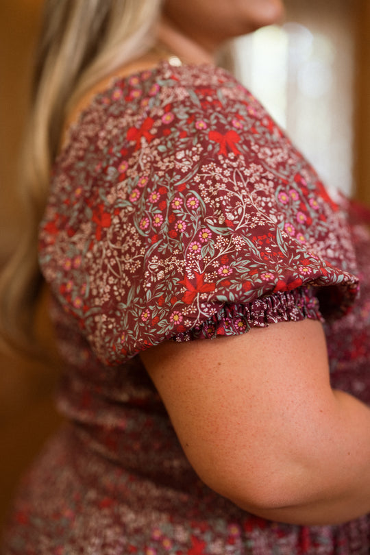 A close-up of a person wearing the Ivy City Co Sunday Best Dress in Garnet Ribboned Garden, featuring short puffed sleeves and a smocked bodice. The softly blurred background and the person’s long hair draped over their shoulder create an elegant holiday dress look.