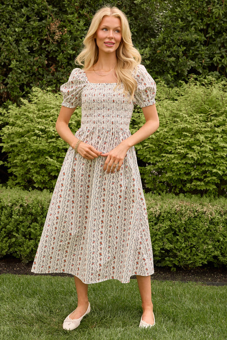 A woman stands outdoors by green bushes, smiling with her hands clasped, wearing the Sunday Best Dress in Linear Poppy Woodblock by Ivy City Co—a light-patterned, short-sleeve maternity midi dress with a square neckline and cream flats.
