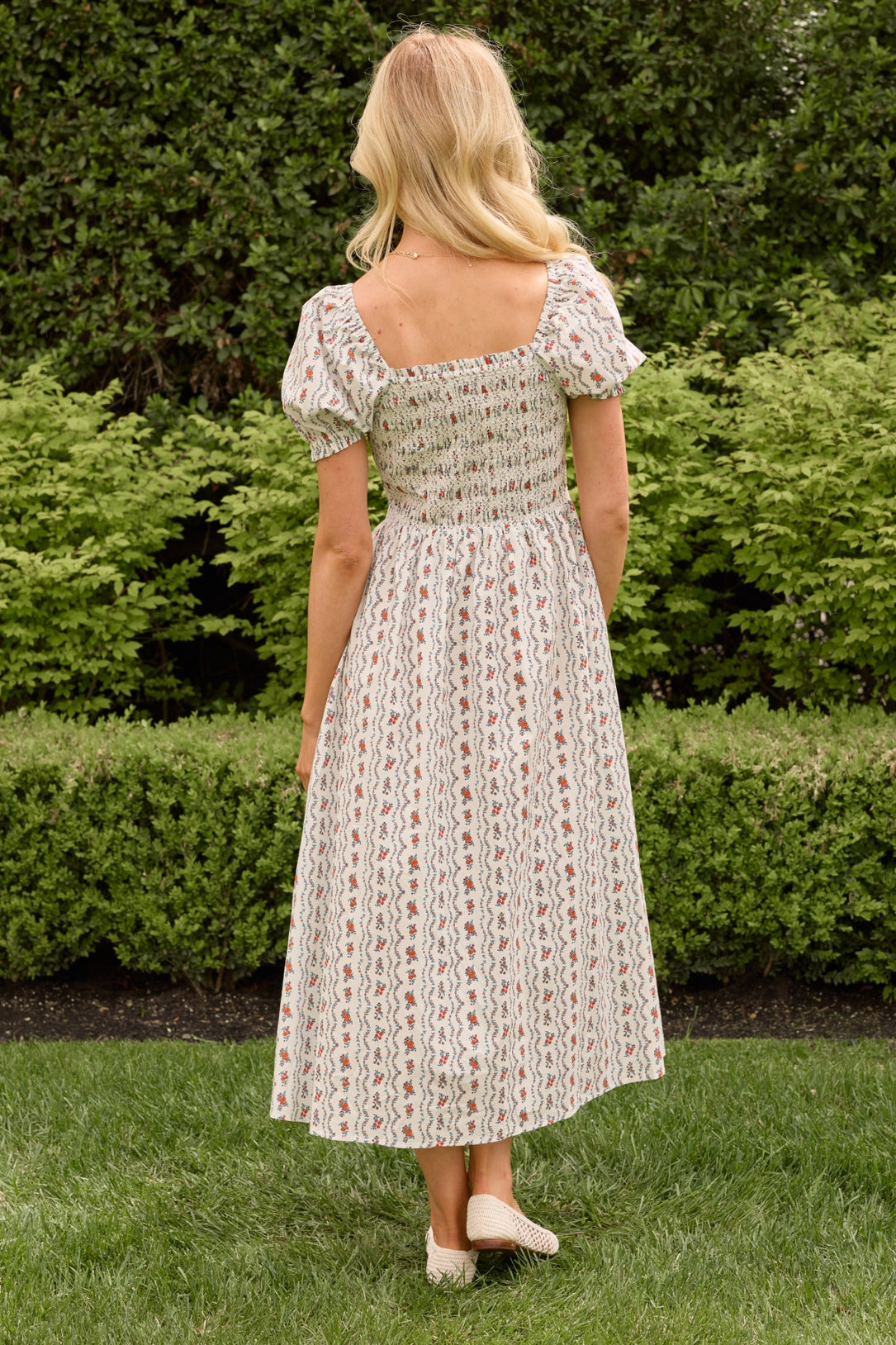 A woman with blonde hair stands outdoors on grass, facing away, wearing the Ivy City Co Sunday Best Dress in Linear Poppy Woodblock. The white midi dress features patterns and puffed sleeves, set against green bushes and trees.