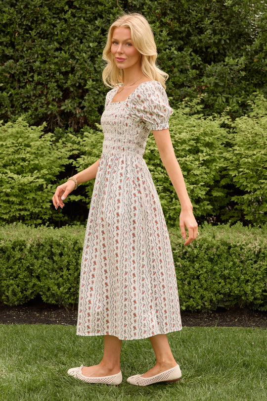 A woman with long blonde hair wears the Ivy City Co Sunday Best Dress in Linear Poppy Woodblock and beige flats, standing on grass before green bushes, looking to the side with a gentle smile.