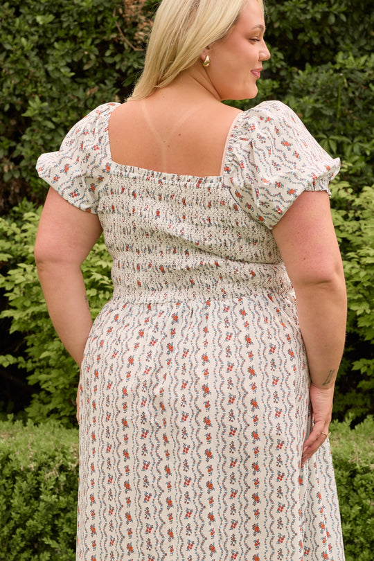 A woman with blonde hair, wearing the Ivy City Co Sunday Best Dress in Linear Poppy Woodblock, stands outdoors facing away from the camera. Lush green shrubs and trees fill the background.