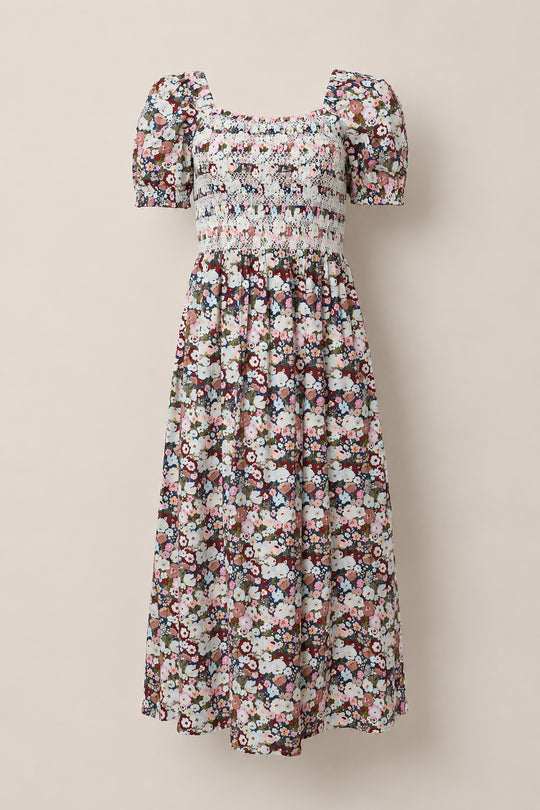 The Ivy City Co Sunday Best Dress in Midnight Garden features puff sleeves and an ankle-length silhouette with a smocked bodice, showcasing a multicolored floral pattern on a white background. Displayed against a neutral backdrop, this maternity-friendly style ensures effortless comfort and elegance.