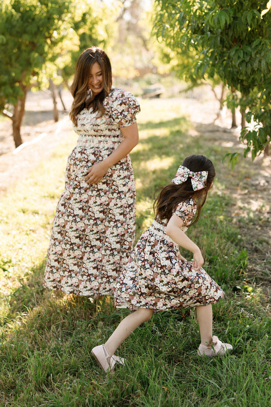A woman and a young girl, both dressed in matching Ivy City Co Sunday Best Dress in Midnight Garden with smocked bodices, enjoy a sunny day outdoors. The woman smiles as the girl runs ahead through the grass among green trees—their maternity-friendly outfits perfect for every joyful moment together.
