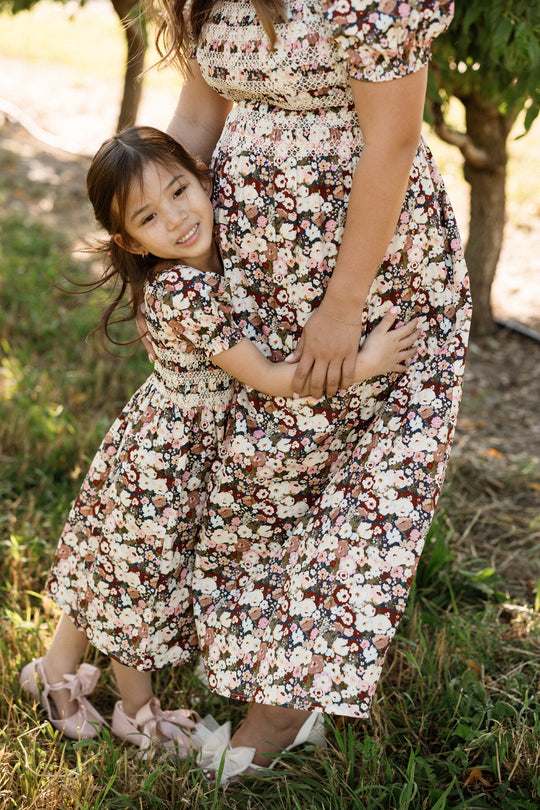 A young girl hugs a woman’s waist outdoors, both wearing matching Ivy City Co Sunday Best Dresses in the Midnight Garden print with smocked bodices and pink shoes. They stand on grass surrounded by trees and sunlight, their maternity-friendly outfits perfect for special moments together.