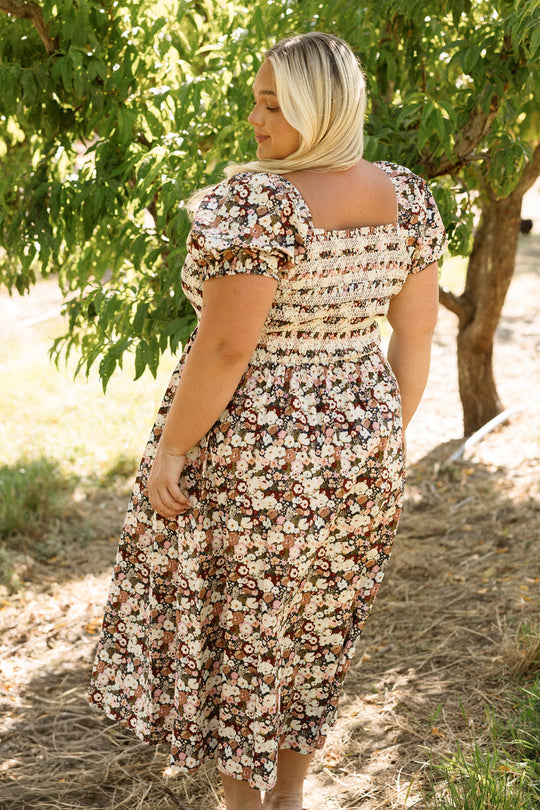 A person in the Ivy City Co Sunday Best Dress in Midnight Garden, featuring a smocked bodice, stands outdoors on a sunny day with their back to the camera. Green trees and grass fill the background, creating an effortlessly chic and maternity-friendly scene.
