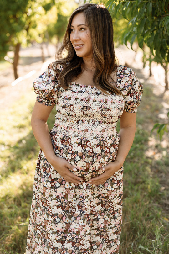 Wearing the Ivy City Co Sunday Best Dress in Midnight Garden, a woman stands outdoors in sunlight, smiling as she cradles her pregnant belly with both hands. Surrounded by trees and greenery, she showcases the dress’s maternity-friendly design.