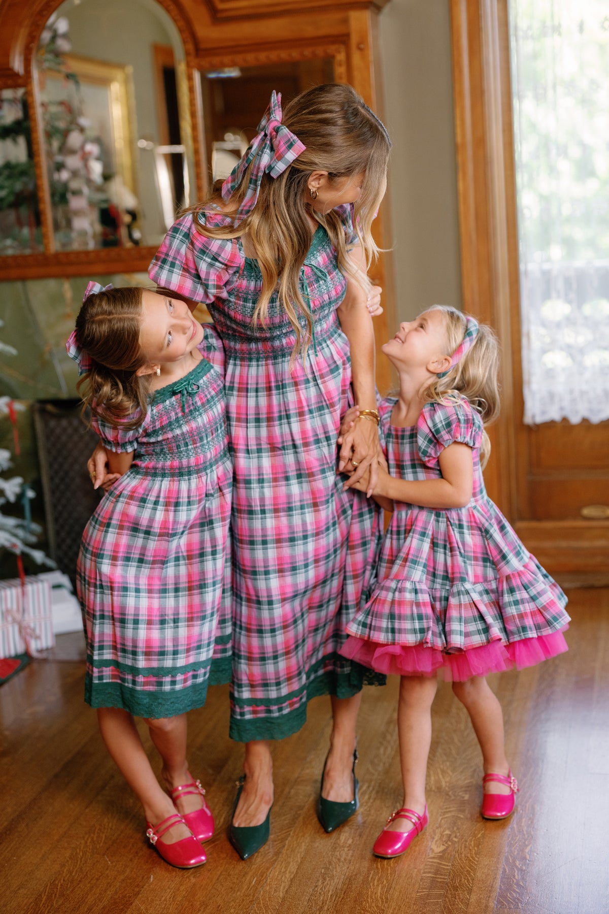 Three generations—one woman and two young girls—smile together indoors by a window and a festive Christmas tree, all dressed in Ivy City Co’s Sunday Best Dress in Vibrant Plaid, featuring matching smocked bodices.