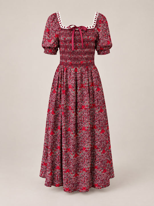 The Sunday Best Dress in Garnet Ribboned Garden by Ivy City Co is a long holiday dress featuring a floral print, puffed short sleeves, a smocked bodice, and a square neckline adorned with white lace and finished with a burgundy ribbon bow at the center.