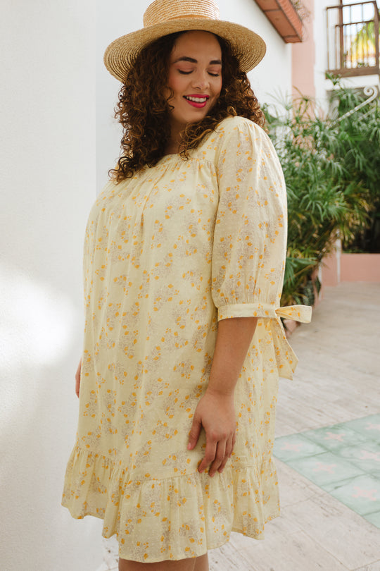 A woman with curly hair wears the Ivy City Co Sunny Dress in Yellow Floral, a maternity-friendly style with a ruffled hem, paired with a straw hat. She smiles outdoors amid lush greenery and a white wall.